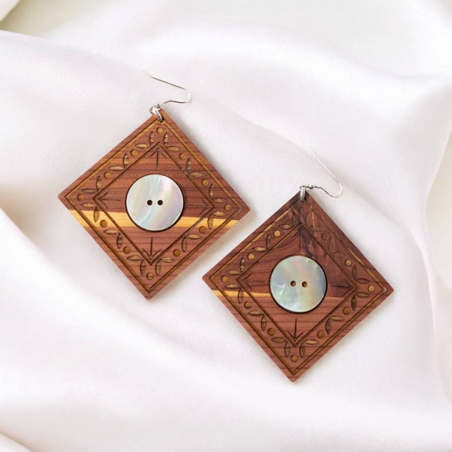 A pair of "Ancestral Framing" earrings in cedar wood from Copper Canoe Woman lay on white fabric.