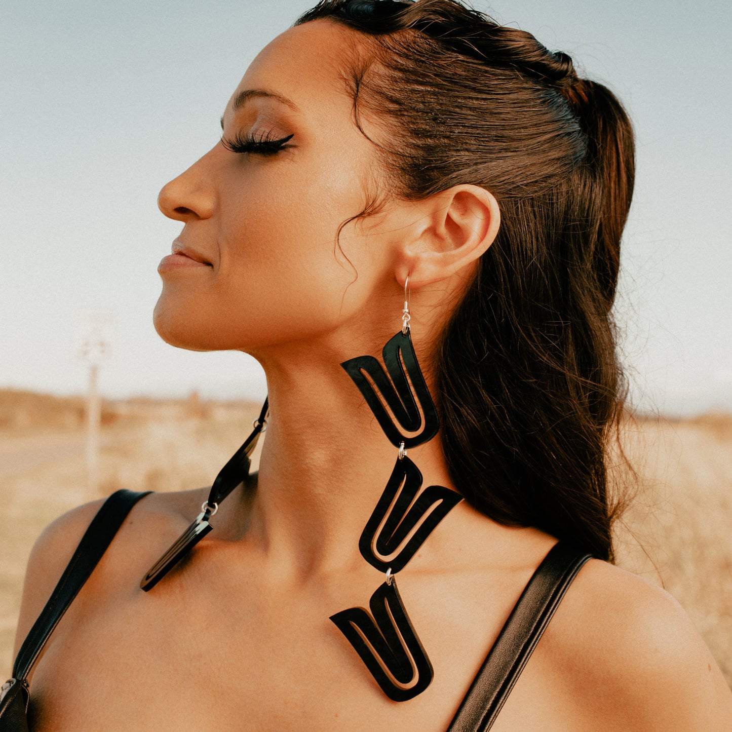 Model stands in field in profile, wearing black acrylic style of the "Formline Cascade" earring from Copper Canoe Woman.