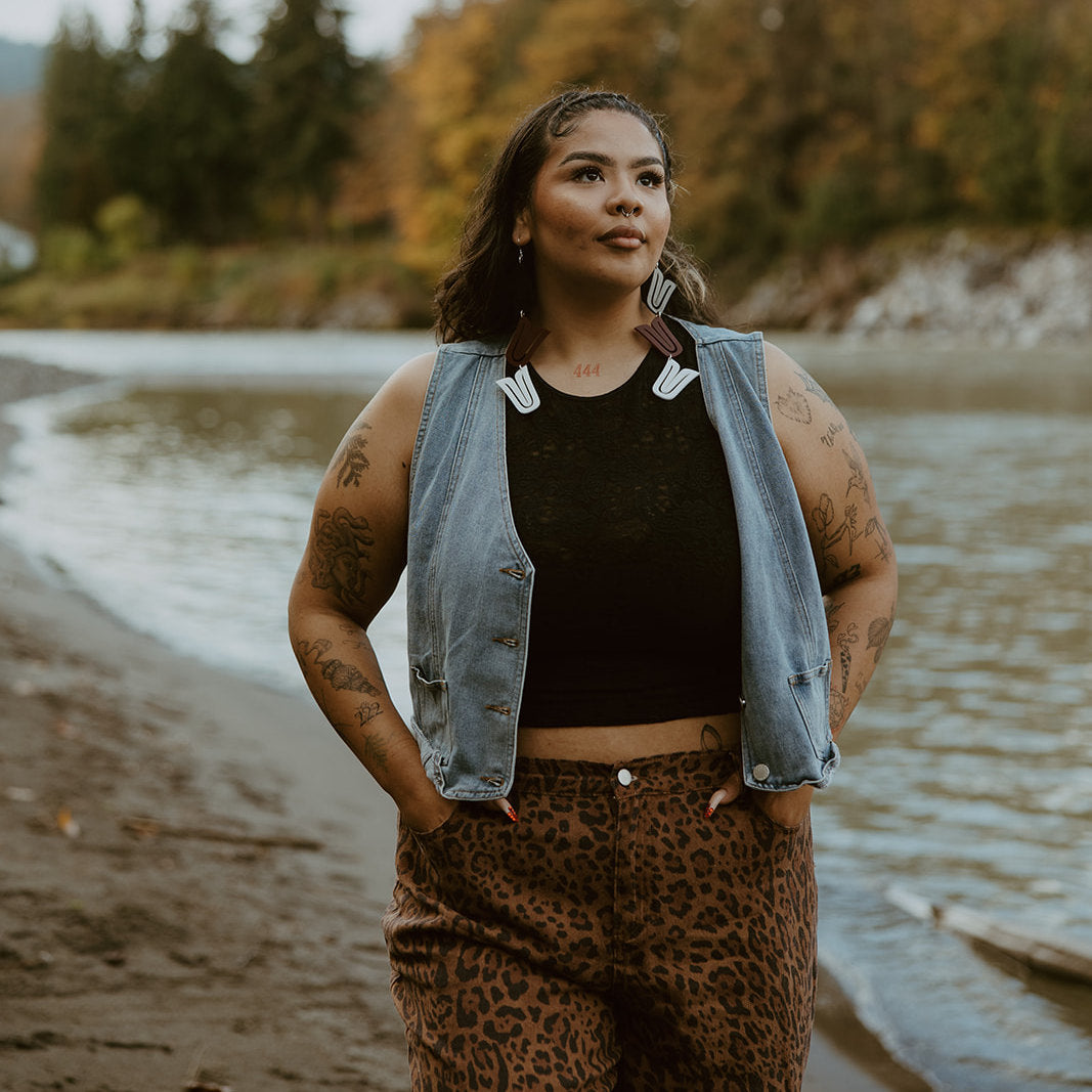 A model stands near a river looking at the sky while wearing a pair of mixed acrylic "Formline Cascade" earrings from Copper Canoe Woman.