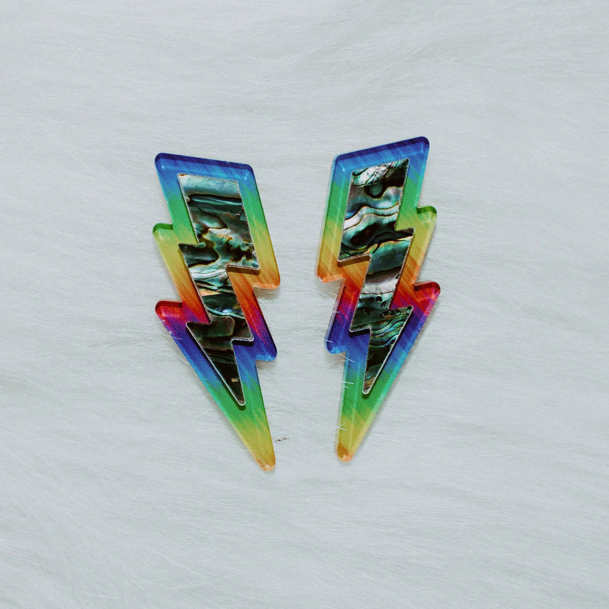 A pair of lightning bolt earrings from Copper Canoe Woman laying on white fur in pride rainbow acrylic with abalone inlay.