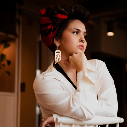 A person wearing a white shirt and a red headpiece wearing earrings from Copper Canoe Woman featuring an eagle wing and a raven wing design.
