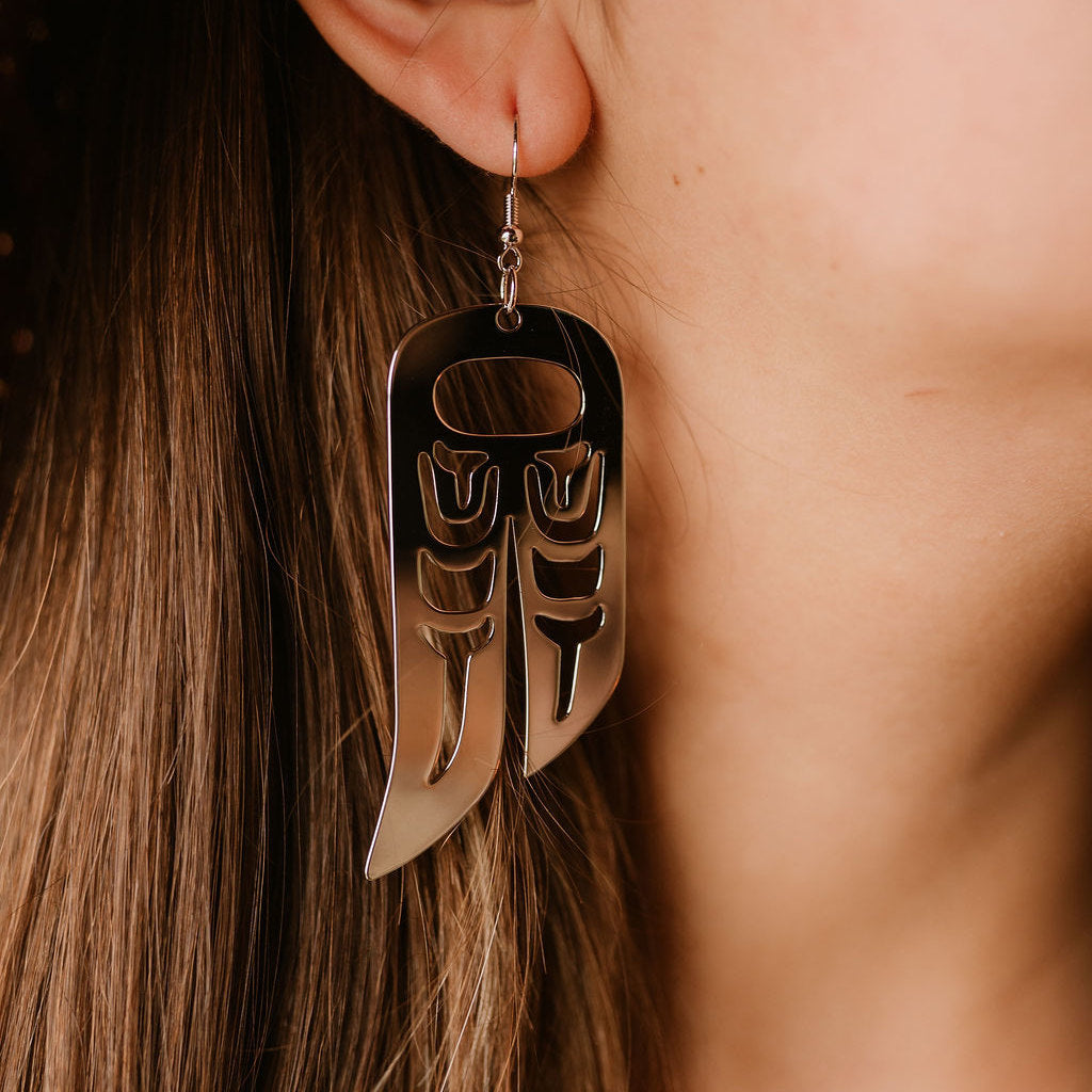 Close-up of a person wearing a rhodium platinum plated earring in the shape of a raven wing using formline Indigenous art.
