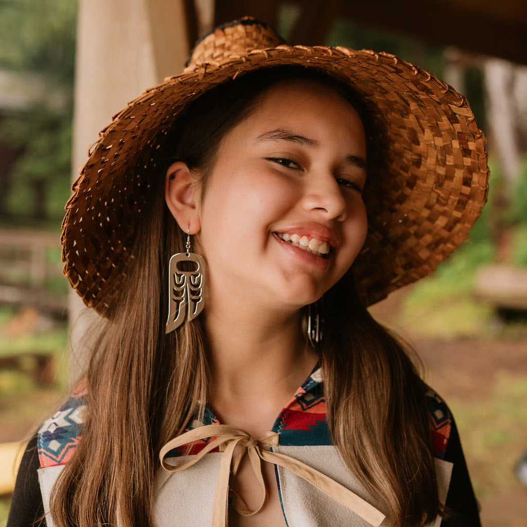 A young woman wearing a cedar hat smile at the camera while rhodium platinum Lovebird wing earrings from Copper Canoe Woman.
