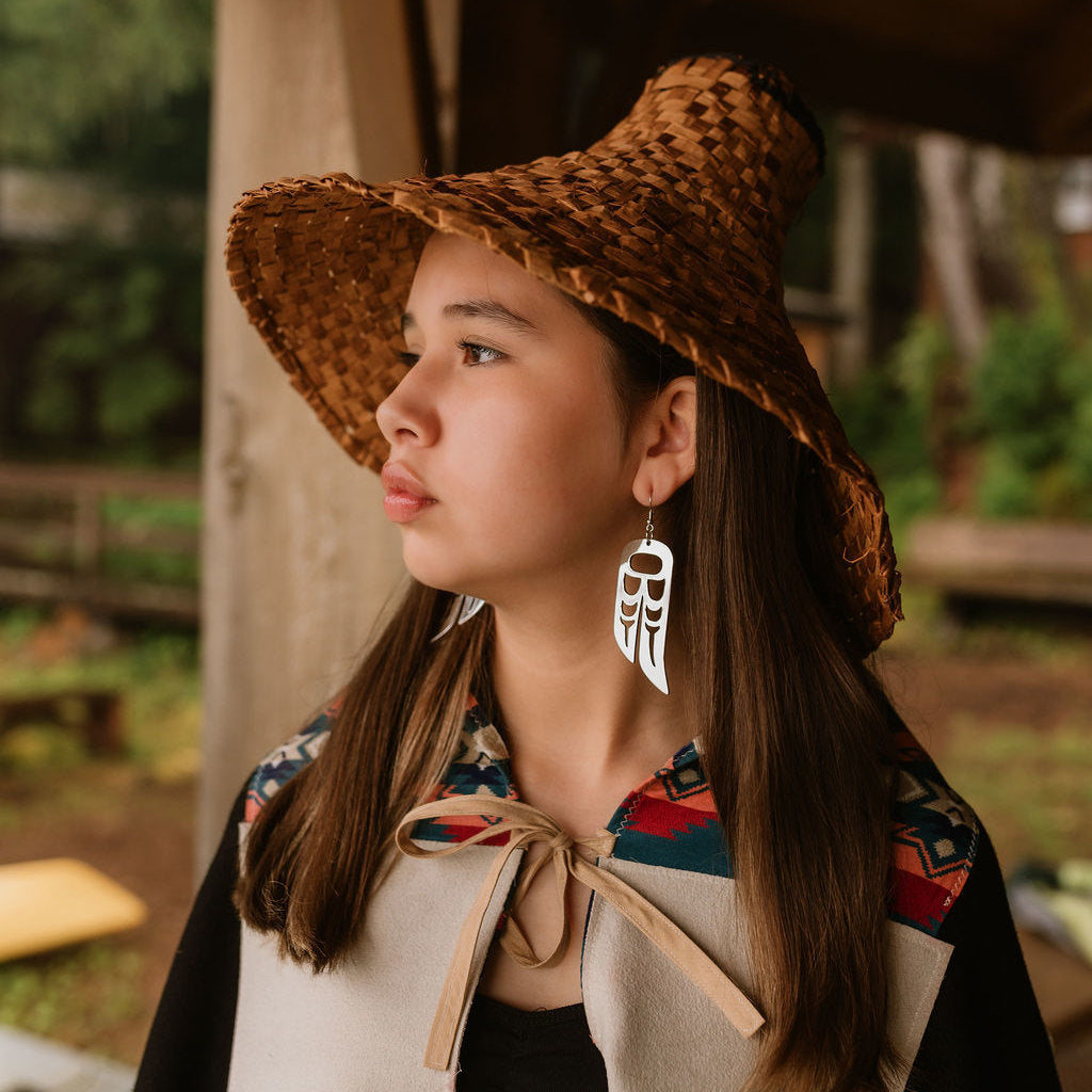 Profile of a young person wearing a cedar hat and rhodium platinum plated Lovebird wing earrings from Copper Canoe Woman.