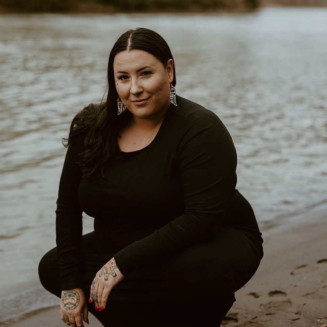 A model kneels by water while wearing silver mirror "Love Birds" earrings from Copper Canoe Woman.