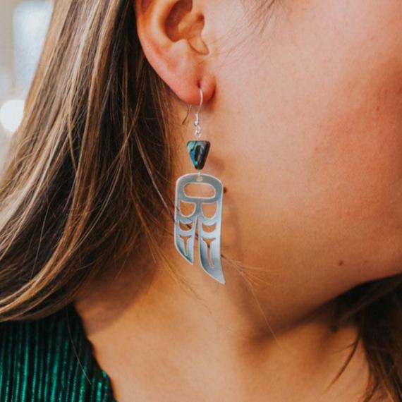 close up of woman wearing Sterling Silver earrings of wings in formline art: one eagle, one raven. Topped with triangle abalone beads from copper canoe woman
