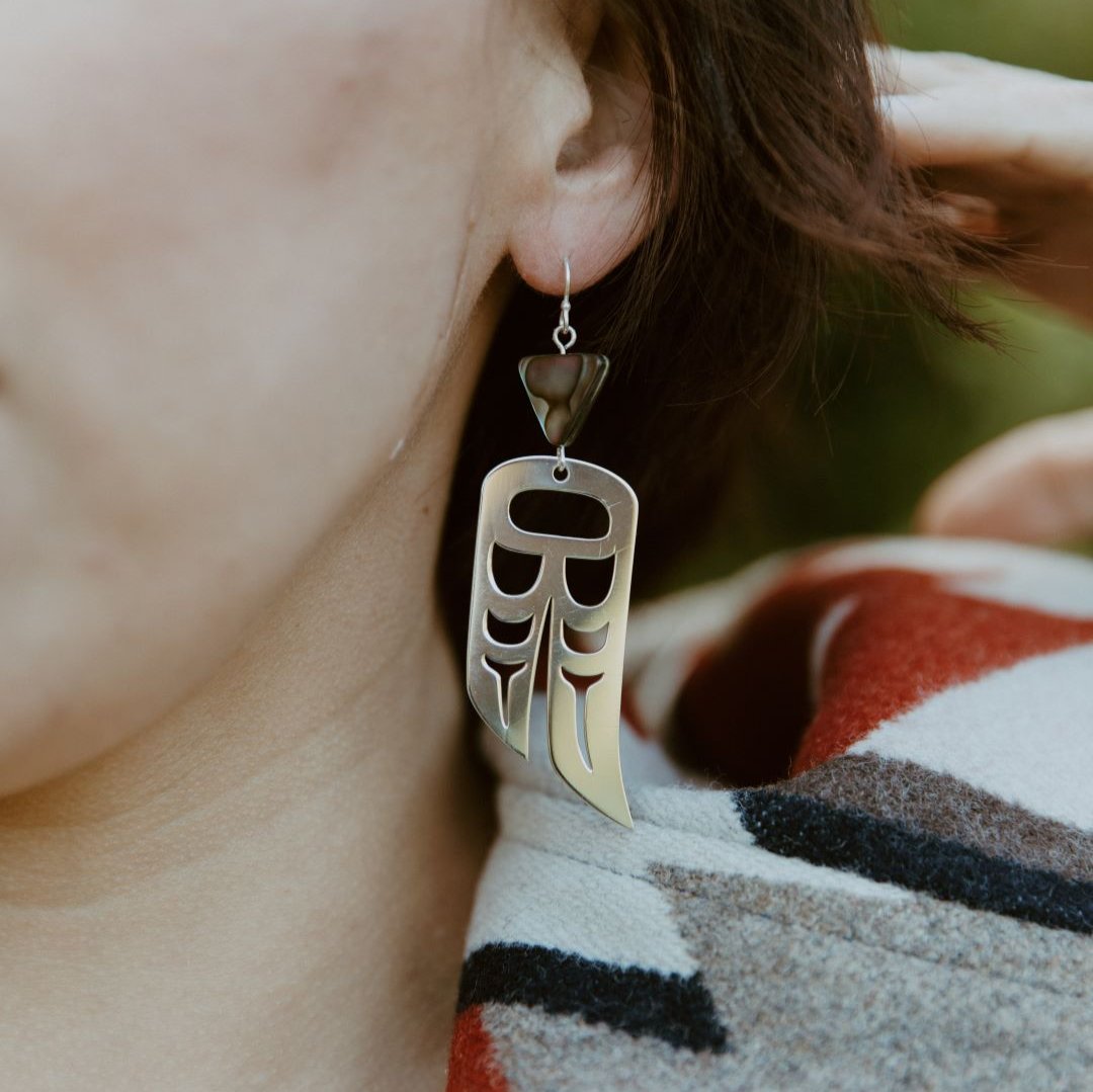 close up of woman wearing Sterling Silver earrings of wings in formline art: one eagle, one raven. Topped with triangle abalone beads from copper canoe woman