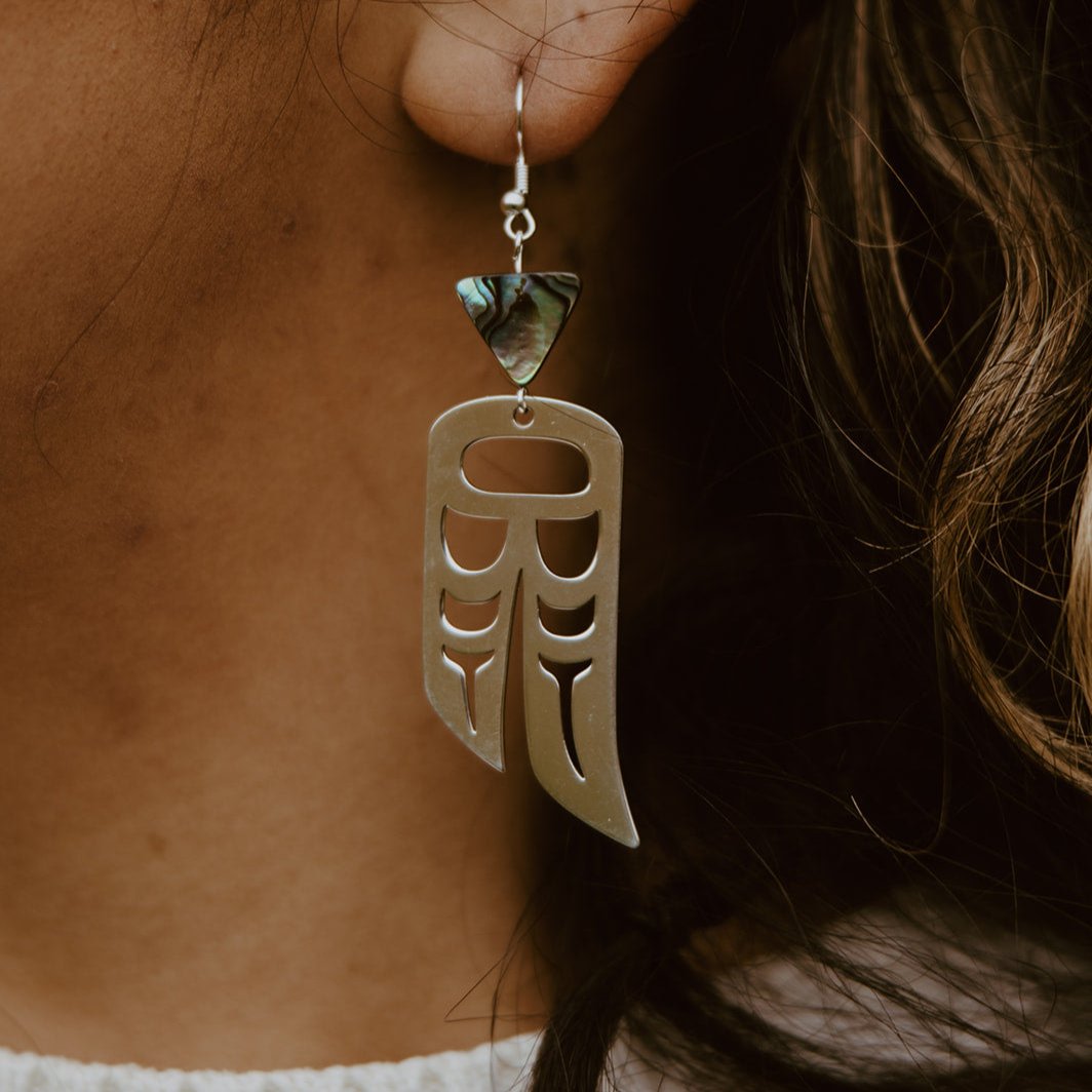 close up of woman wearing Sterling Silver earrings of wings in formline art: one eagle, one raven. Topped with triangle abalone beads from copper canoe woman