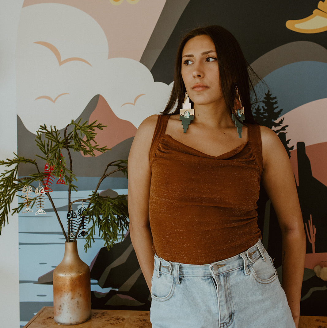A model stands in front of a mural wearing gold mirror and sage acrylic "Mesa" earrings from Copper Canoe Woman.