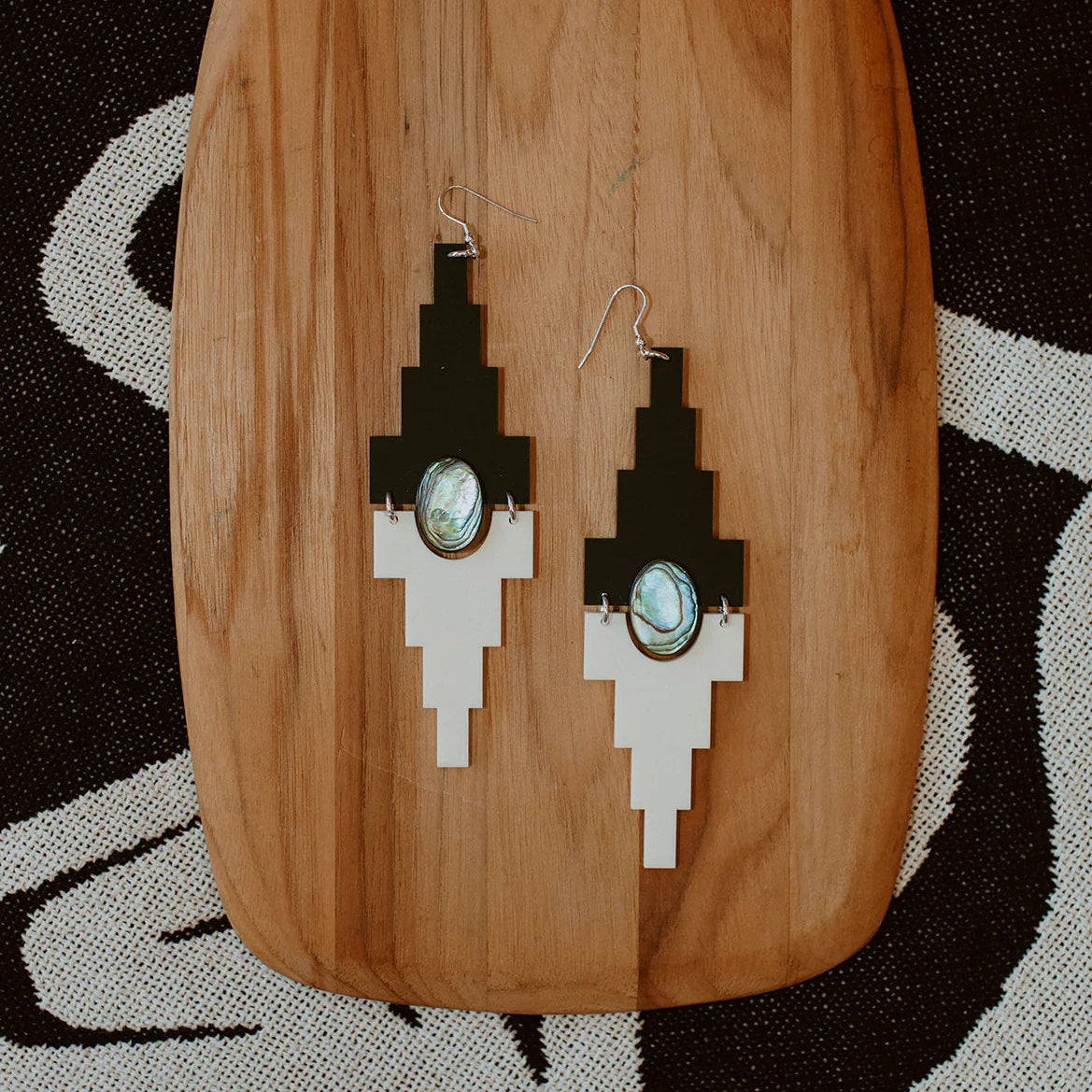 A pair of jet black and cream acrylic "Mesa" earrings lay on a wood plank from Copper Canoe Woman.
