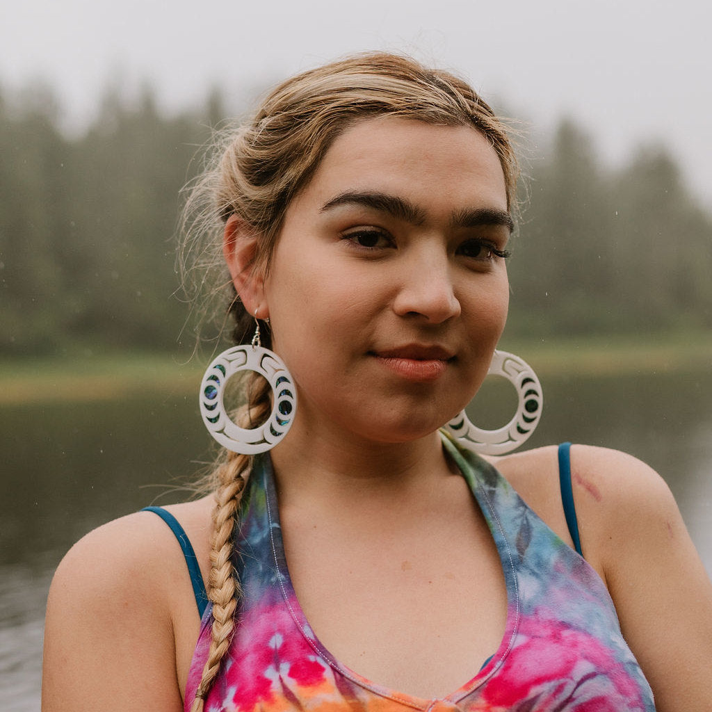 A model stares at the camera near the water wearing white acrylic Nala Hoop earrings with abalone in lay from Copper Canoe Woman.