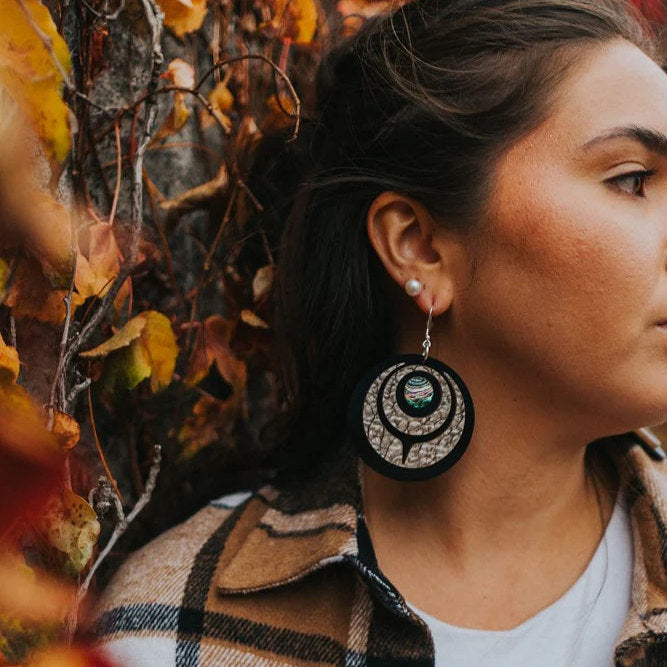 The desert pattern acrylic "Owl Eye" earring from Copper Canoe Woman worn by a model standing in front of leaves.
