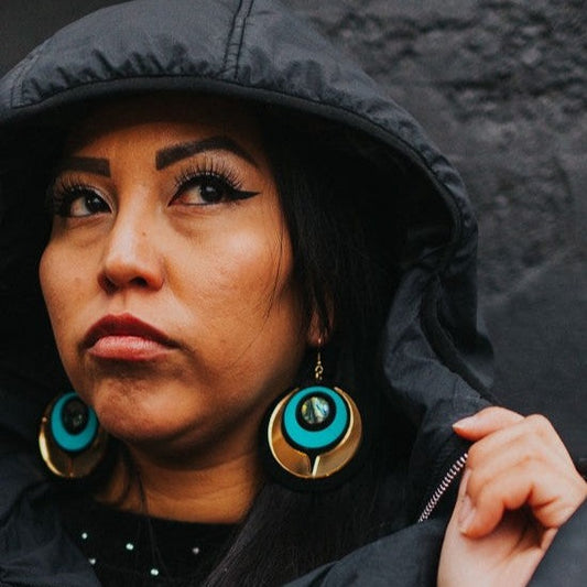 A woman wearing Copper Canoe Woman "Owl Eye" earrings in turquoise and gold mirror acrylic.