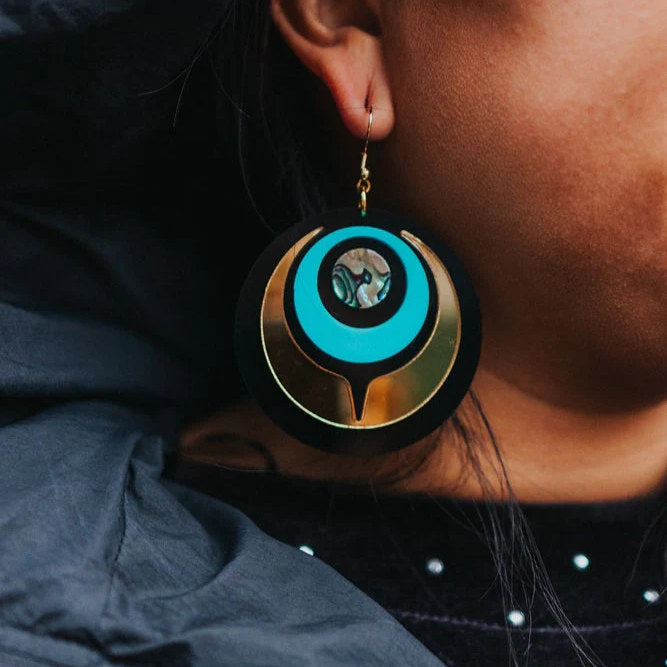 A closeup of the Copper Canoe Woman turquoise and gold mirror acrylic "Owl Eye" earring worn by a model.