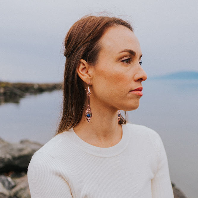 A woman at a beach wearing rose gold mirror acrylic "Paddle Song" earrings from Copper Canoe Woman.