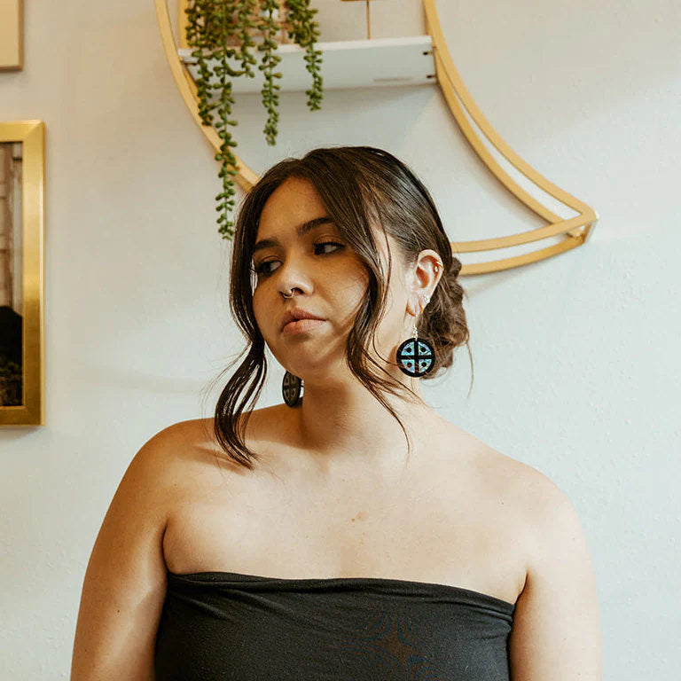 A model stands wearing black acrylic "Protector Shield" earrings with abalone inlay from Copper Canoe Woman.