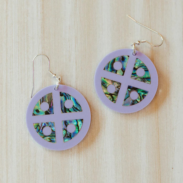 Close up of Copper Canoe Woman "Protector Shield" earrings in lavender acrylic with abalone inlay sitting on a table.