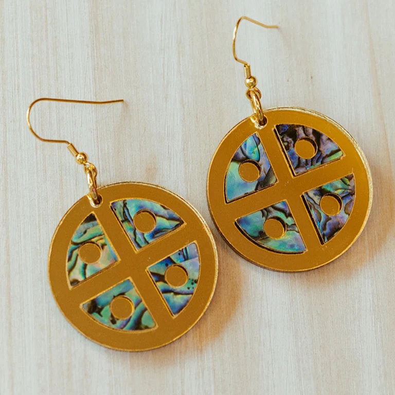 A pair of gold mirror acrylic "Protector Shield" earrings from Copper Canoe Woman rest on a wood table.