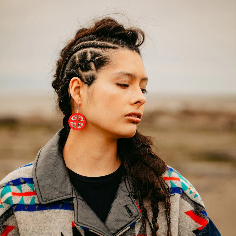 A model standing in a field wears "Protector Shield" earrings in red mirror acrylic from Copper Canoe Woman.