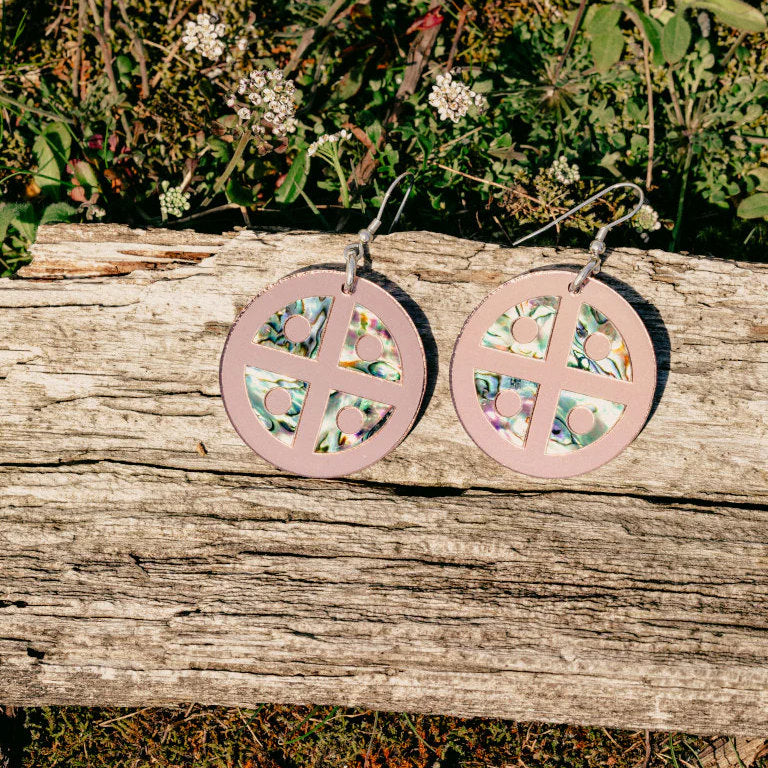 A pair of rose gold mirror acrylic "Protector Shield" earrings from Copper Canoe Woman sit on a log.