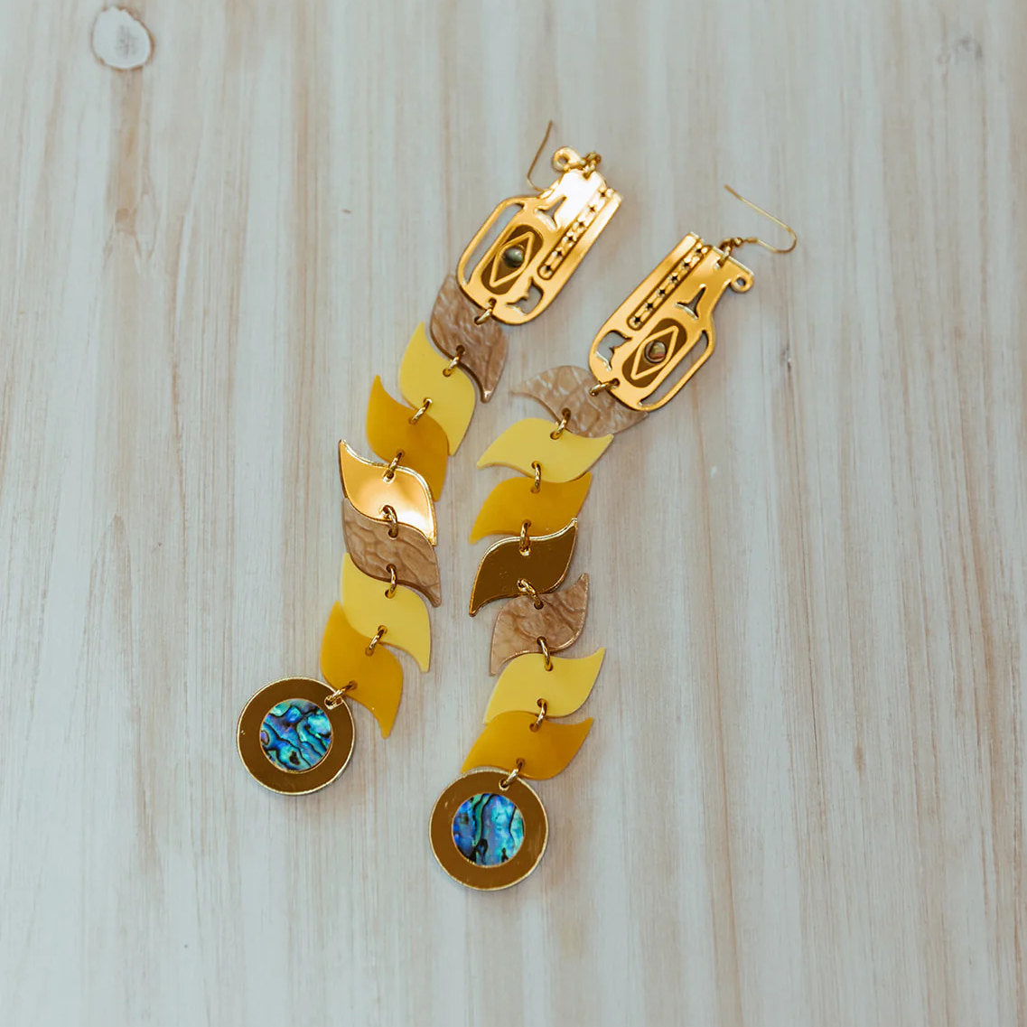 A pair of serpent earrings in yellow and gold mirror acrylic laying on a wooden panel from Copper Canoe Woman.
