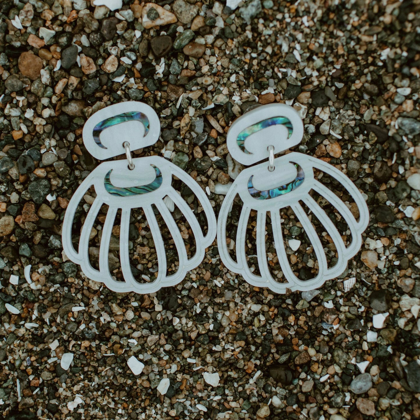 A pair of pearl acrylic "Shell Woman" earrings from Copper Canoe Woman rest on pebbles.