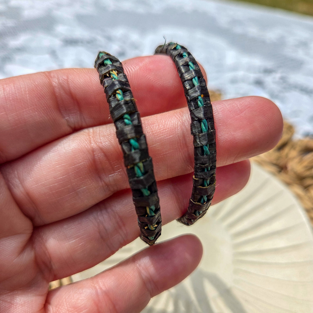 Two black and green patterned bracelets held between fingers with a blurred outdoor background