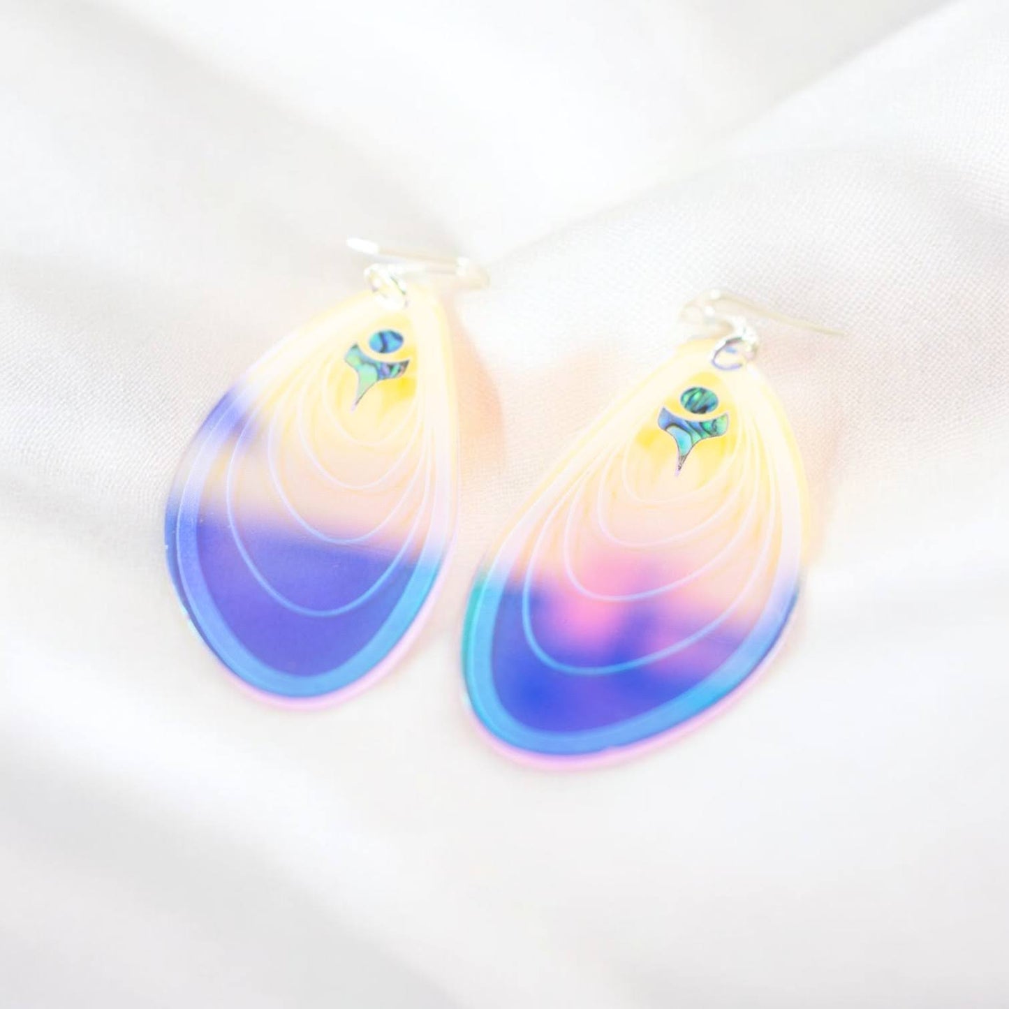 A pair of iridescent acrylic "Tidal Ripples" earrings from Copper Canoe Woman lay on a bed of white fabric.