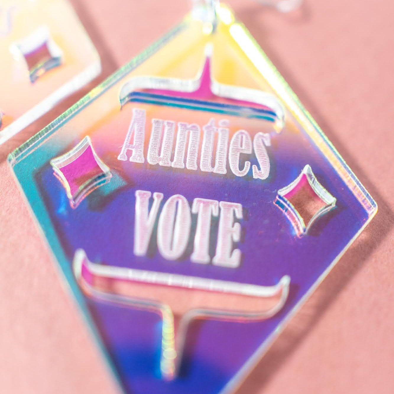 close up of Diamond shaped iridescent earrings with formline cut out and 'Aunties Vote' engraved in the middle from copper canoe woman