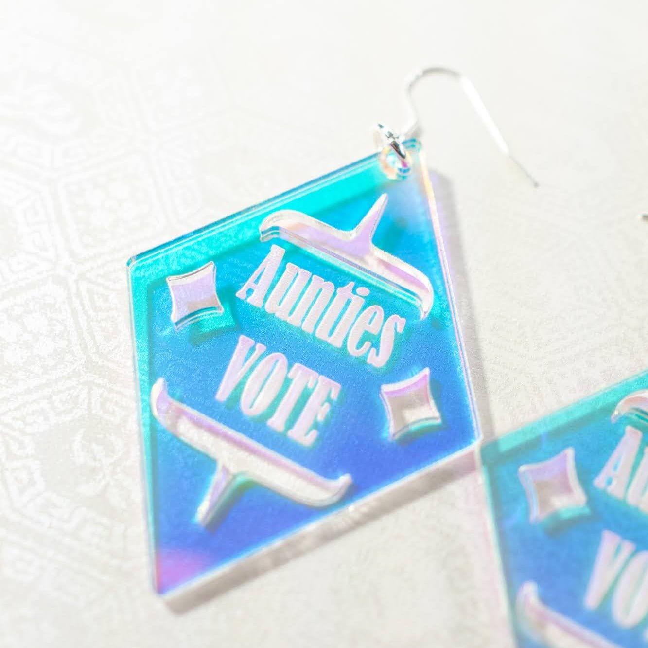 close up of Diamond shaped iridescent earrings with formline cut out and 'Aunties Vote' engraved in the middle from copper canoe woman