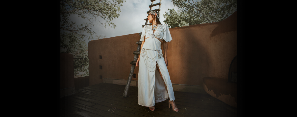 Woman in a white outfit standing in front of a textured wall with a ladder and trees in the background.