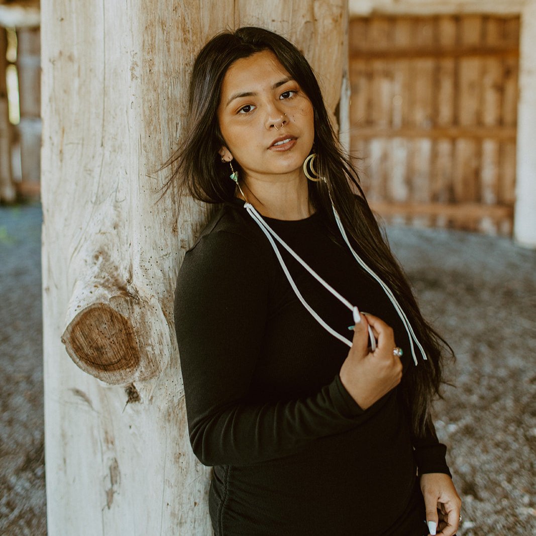 woman wearing Brass crescent moon earrings topped with triangle abalone beads and white fringe hide that hangs from bottom of moon. Designed by Copper Canoe Woman