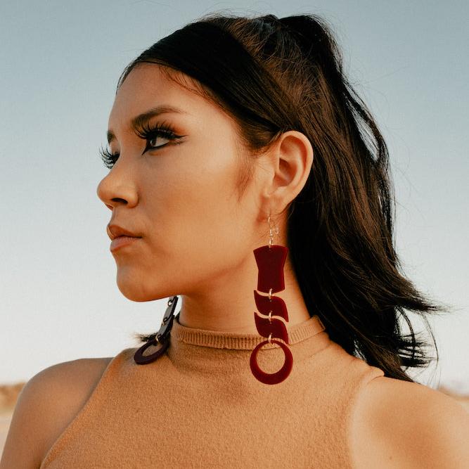 Woman wearing abstract formline art in maroon color. Top is copper shield, followed by two s shaped formline shapes, and the bottom a circle. earrings from copper canoe woman
