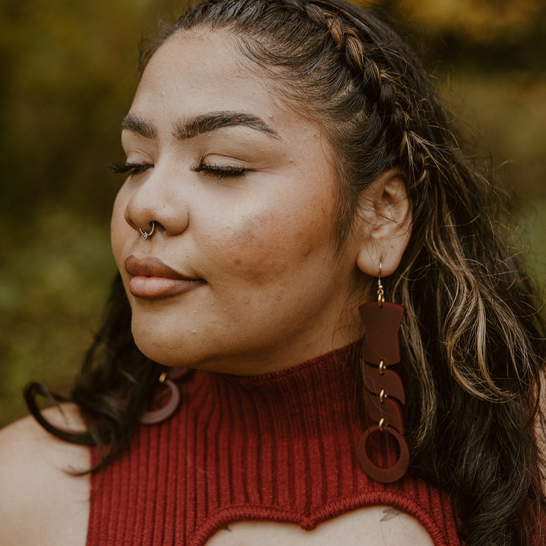 Woman wearing abstract formline art in maroon color. Top is copper shield, followed by two s shaped formline shapes, and the bottom a circle. earrings from copper canoe woman