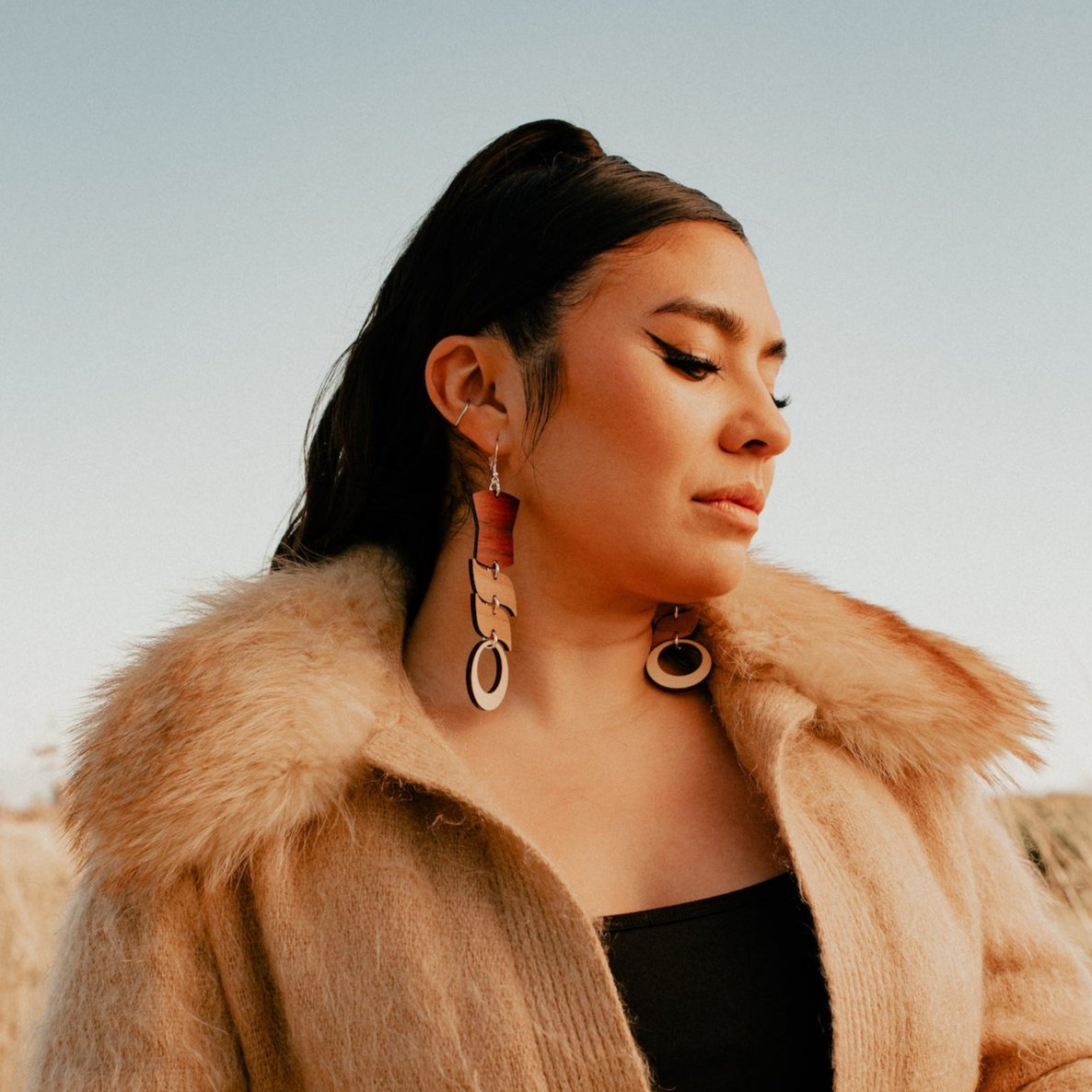 Woman wearing abstract formline art in wood variations. Top is copper shield, followed by two s shaped formline shapes, and the bottom a circle. earrings from copper canoe woman