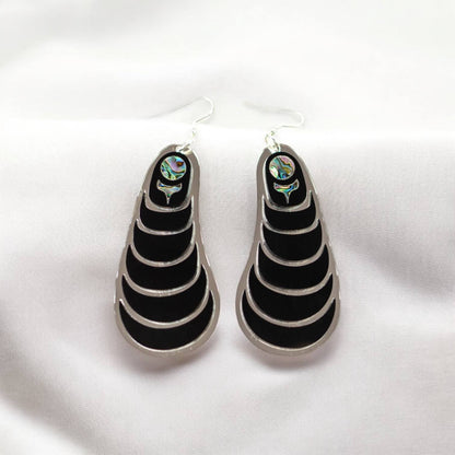 A pair of Copper Canoe Woman "Coastal Strata" earrings in black acrylic laying on white fabric.