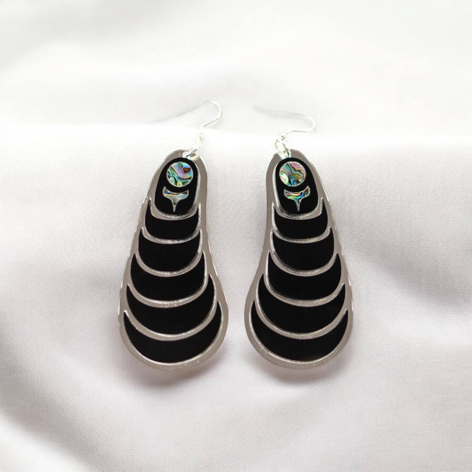 A pair of Copper Canoe Woman "Coastal Strata" earrings in black acrylic laying on white fabric.