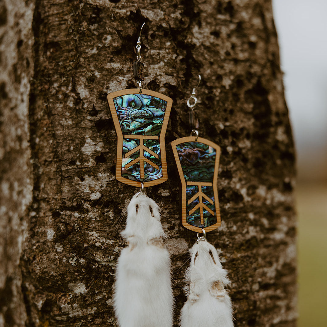 A closeup of copper shield shaped earrings with abalone inlay and ermine pelts hanging from a tree in a field. Design by Copper Canoe Woman.