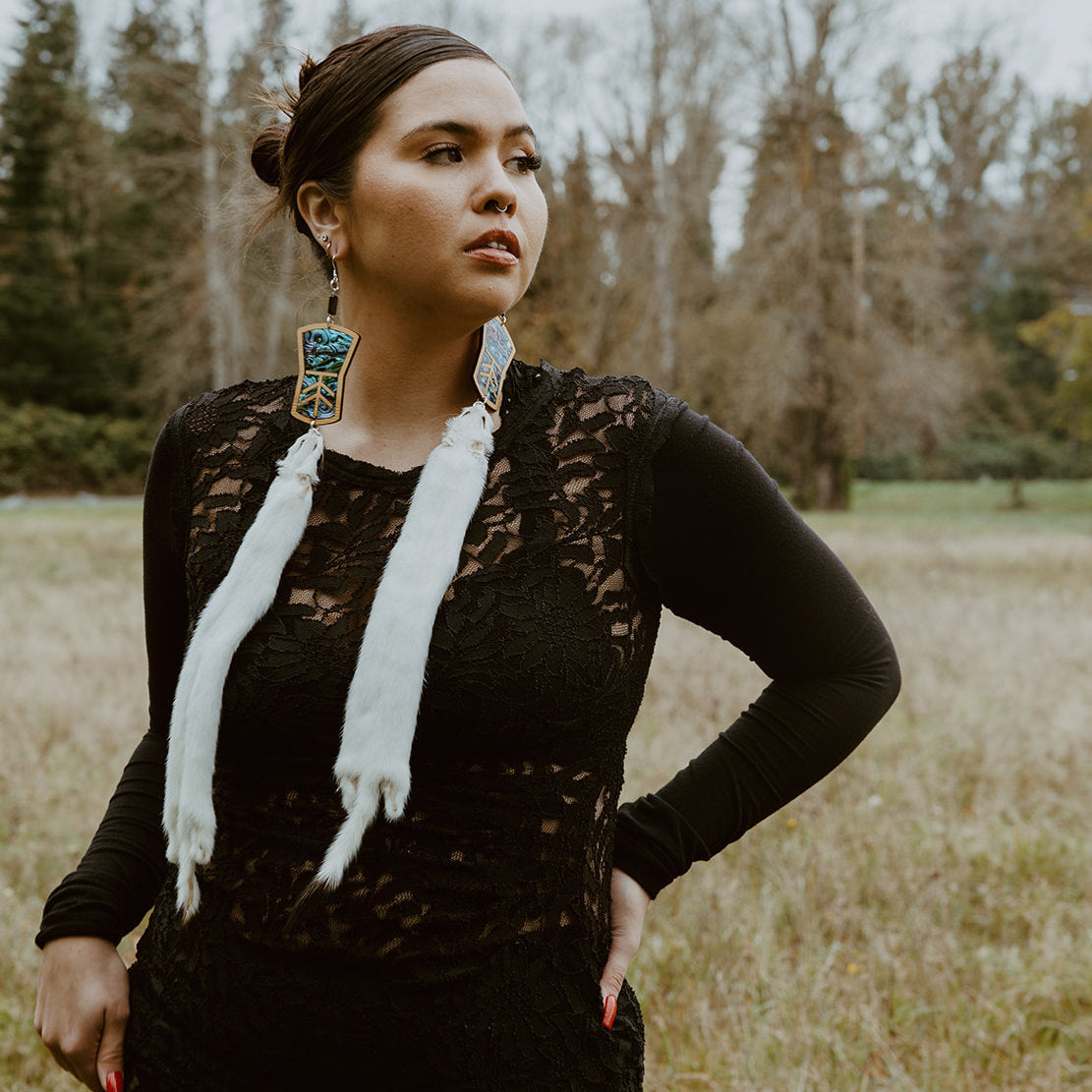 A model stands in a field looking into the distance while wearing copper shield shaped earrings with abalone inlay and ermine pelt. Design by Copper Canoe Woman.