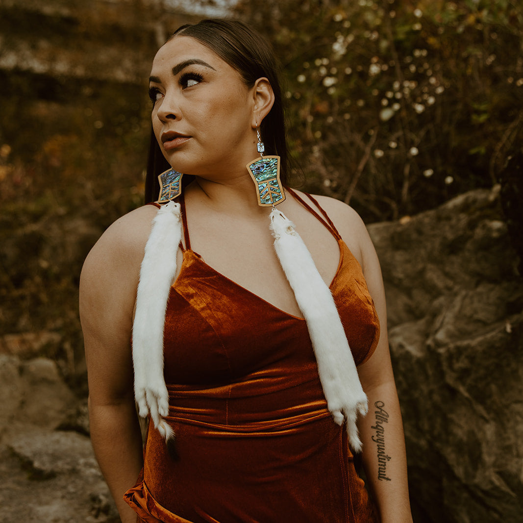 Model looks over her shoulder while wearing copper shield shaped earrings with abalone inlay and ermine pelt. Design by Copper Canoe Woman.
