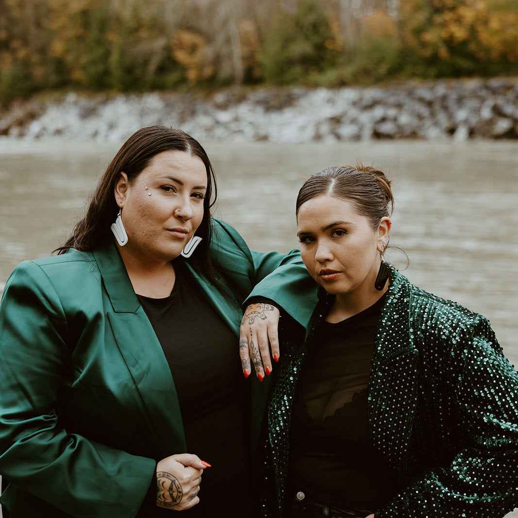 Two models stand together facing the camera while wearing "Echoed U" earrings from Copper Canoe Woman, the left in white acrylic and right in black acrylic.