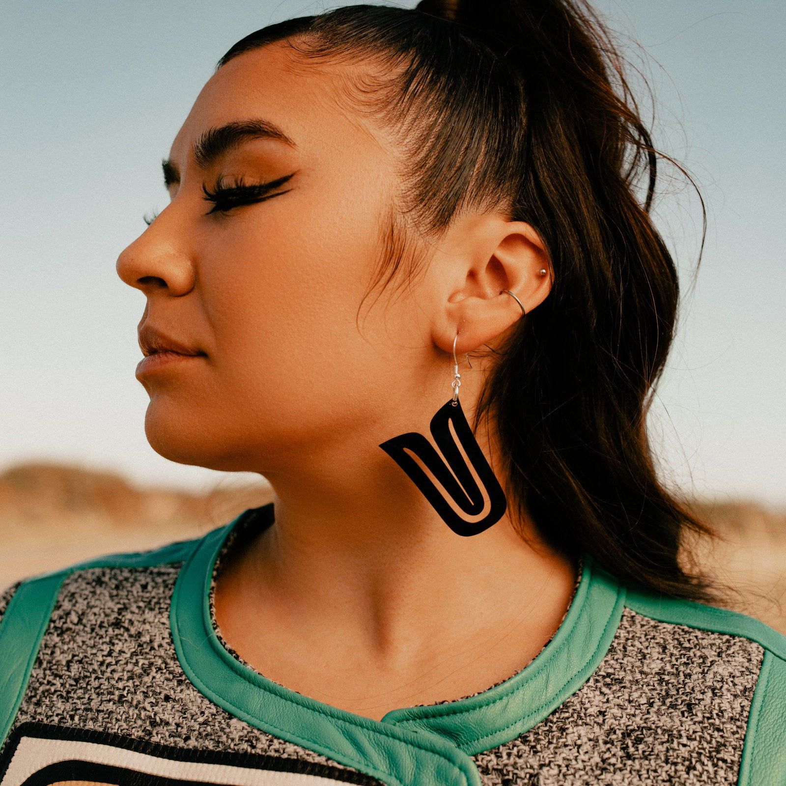 A model wearing a pair of black acrylic "Echoed U" earrings by Copper Canoe Woman stands in a field.