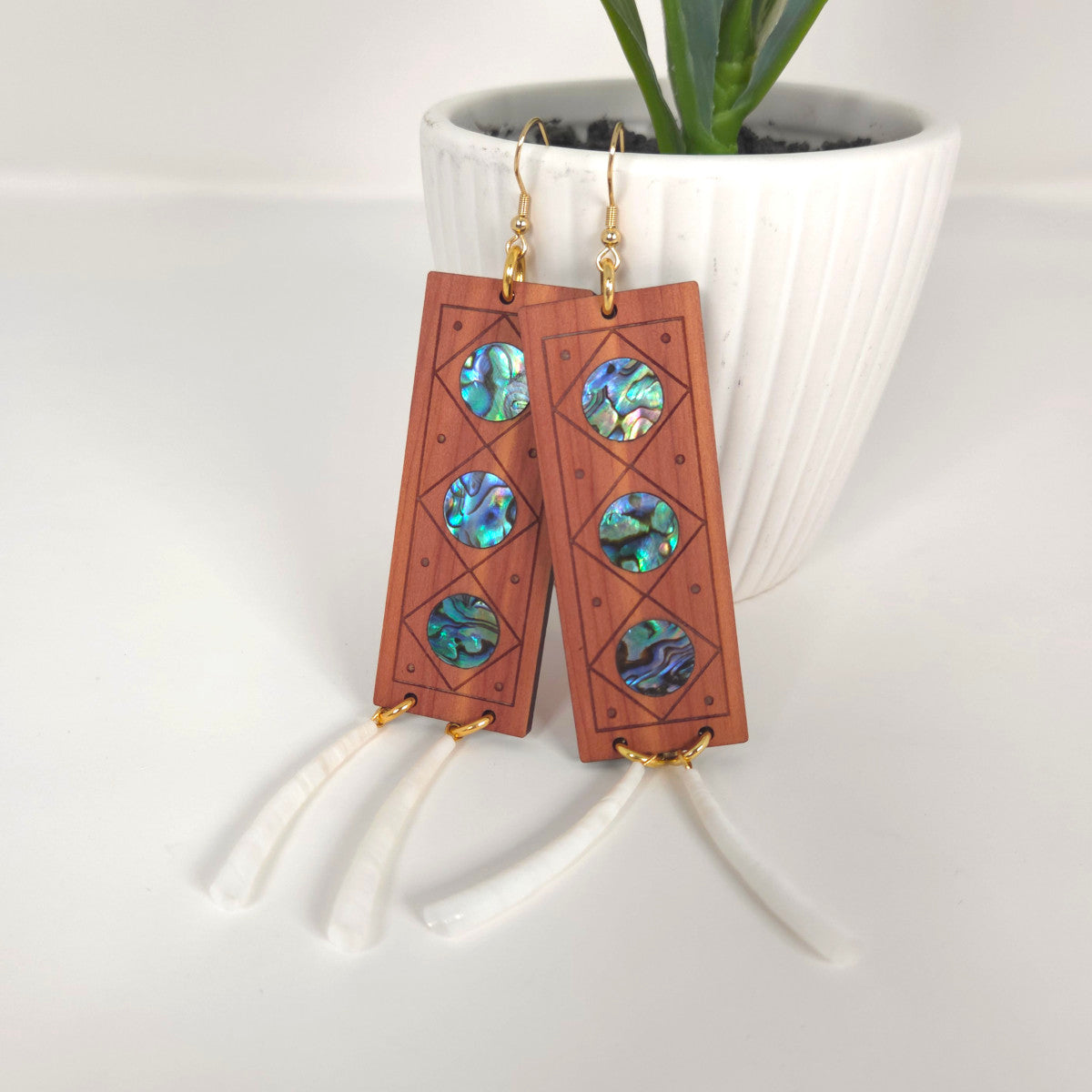 Handmade wooden earrings with abalone shell inlays on a white surface hang from a potted plant.