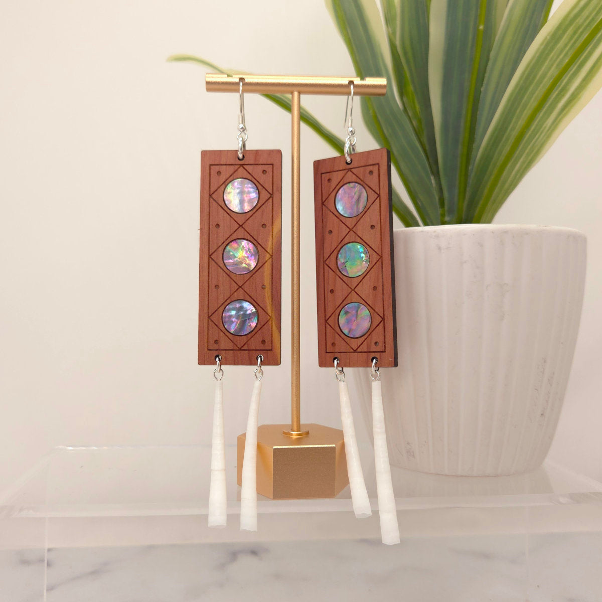 Wooden earrings with mother of pearl inlay on a gold stand with a plant in the background.