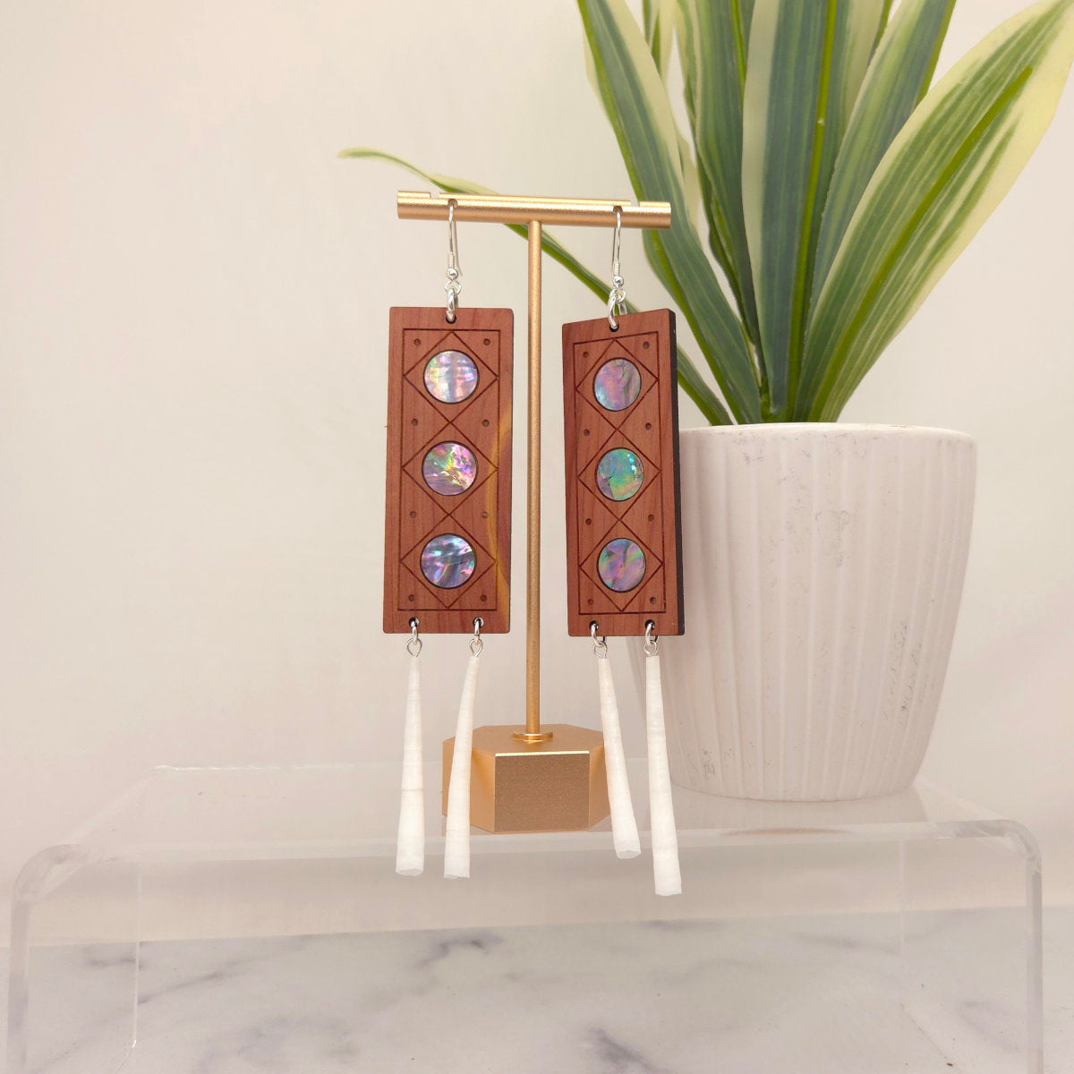 Handmade cedar wood earrings with mother of pearl inlay and dentalium shell on a gold stand, with a plant in the background.