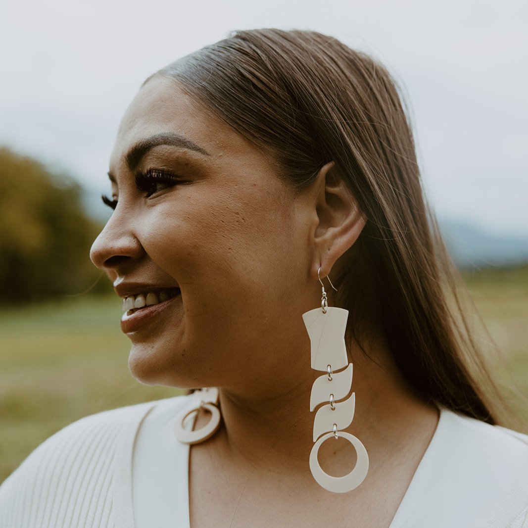 Woman wearing abstract formline art in ivory color. Top is copper shield, followed by two s shaped formline shapes, and the bottom a circle. earrings from copper canoe woman