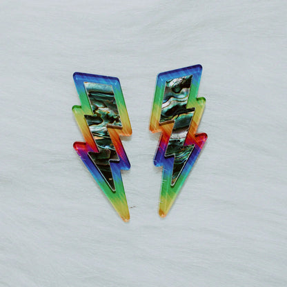 A pair of lightning bolt earrings from Copper Canoe Woman laying on white fur in pride rainbow acrylic with abalone inlay.