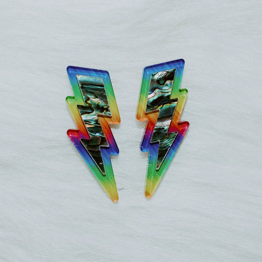 A pair of lightning bolt earrings from Copper Canoe Woman laying on white fur in pride rainbow acrylic with abalone inlay.