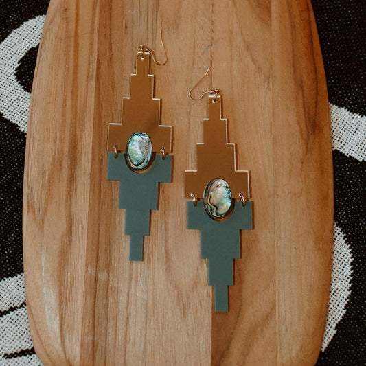 A pair of Mesa earrings from Copper Canoe Woman in gold mirror and sage acrylic lay on a wood plank.