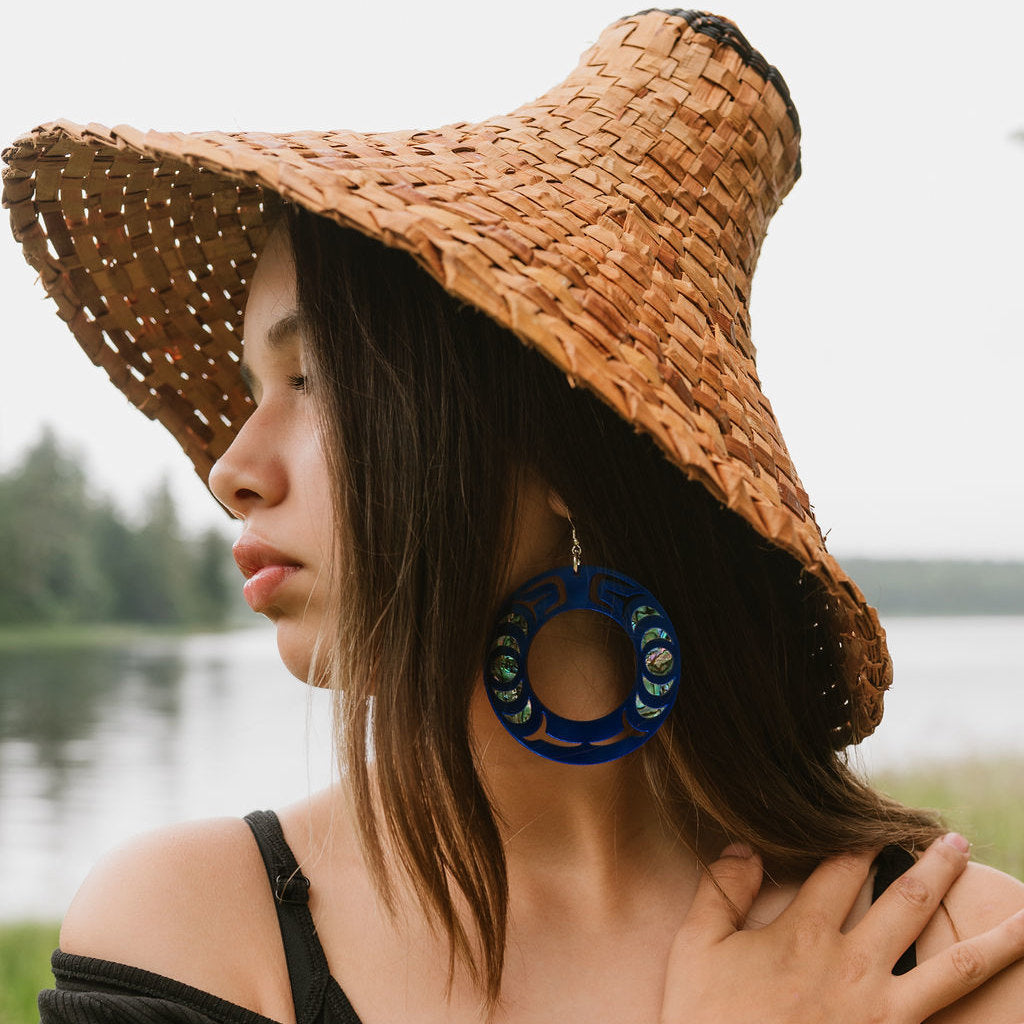 Woman wearing a cedar hat and Copper Canoe Woman cobalt mirror acrylic Nala Hoop earrings in profile by a lake.
